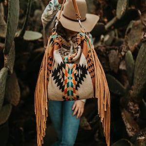 May include: A brown leather shoulder bag with fringe details. The bag is made of a woven fabric with a geometric pattern in shades of orange, blue, and white. The bag is being held by a person in front of a cactus.