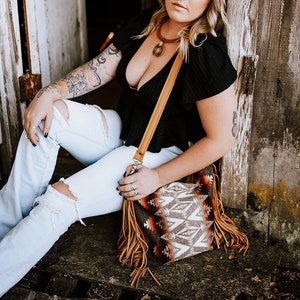 May include: A woman wearing a black top and light blue ripped jeans, sitting next to a wooden door. She is wearing a brown necklace and carrying a brown and patterned shoulder bag with fringe.