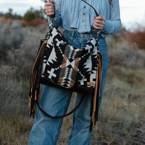Puede incluir: Un bolso de hombro estilo hobo con un patr&oacute;n geom&eacute;trico en negro, blanco y beige. El bolso incluye largos flecos superpuestos en beige y negro a ambos lados, una base negra y una correa de hombro negra sujeta con herrajes de metal plateado.