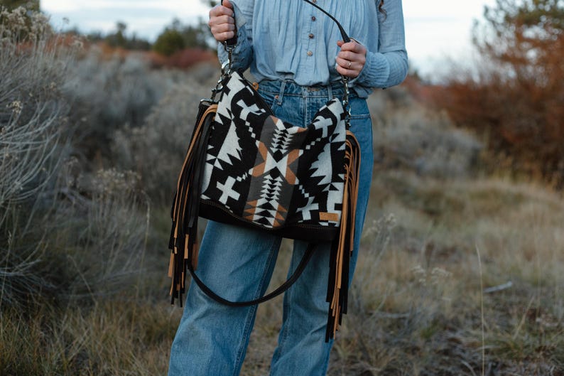 Puede incluir: Un bolso de hombro estilo hobo con un patr&oacute;n geom&eacute;trico en negro, blanco y beige. El bolso incluye largos flecos superpuestos en beige y negro a ambos lados, una base negra y una correa de hombro negra sujeta con herrajes de metal plateado.