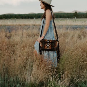 May include: A light blue tiered maxi dress with a subtle floral pattern, paired with a wide-brimmed beige felt hat and a dark brown leather crossbody bag featuring a geometric patterned fabric panel and long fringe accents on the side.
