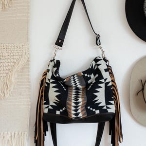 Puede incluir: Un bolso de hombro estilo hobo con un patr&oacute;n geom&eacute;trico en negro, blanco y beige sobre un cuerpo de tela suave. El bolso incluye largos flecos beige en ambos lados, una base negra de aspecto cuero y una correa de hombro negra ajustable.