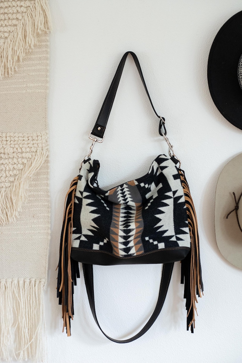 Puede incluir: Un bolso de hombro estilo hobo con un patr&oacute;n geom&eacute;trico en negro, blanco y beige sobre un cuerpo de tela suave. El bolso incluye largos flecos beige en ambos lados, una base negra de aspecto cuero y una correa de hombro negra ajustable.