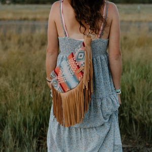 May include: A crossbody bag featuring a vibrant, multi-colored geometric pattern in shades of orange, blue, and white, accented with long, tan-colored leather fringe hanging from the bottom edge and a matching tan leather strap attached to the top.