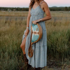 May include: A light blue floral maxi dress with a V-neckline and spaghetti straps, paired with a crescent-shaped crossbody bag featuring a tan leather base and a turquoise fabric panel with a geometric pattern and a long adjustable leather strap.