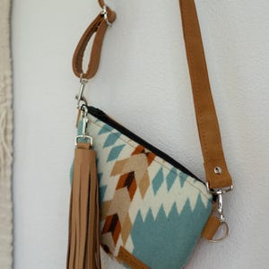 May include: A crossbody bag featuring a light blue, cream, and brown geometric pattern. The bag has a black zipper closure, a long tan strap, and a large tan leather tassel attached to a silver metal clip on the side of the bag.