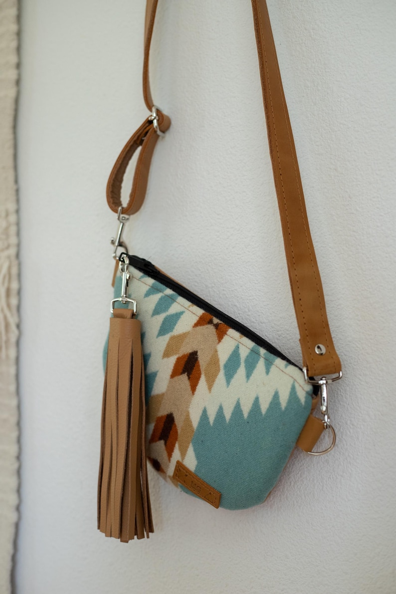 May include: A crossbody bag featuring a light blue, cream, and brown geometric pattern. The bag has a black zipper closure, a long tan strap, and a large tan leather tassel attached to a silver metal clip on the side of the bag.