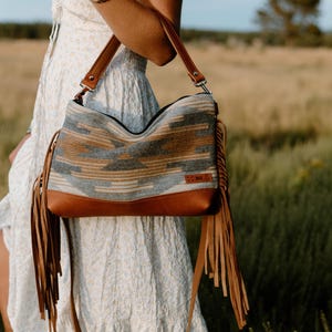 Peut inclure: Un sac à bandoulière doté d'un panneau en tissu tissé avec un motif géométrique dans des tons gris, beige et crème. Le sac possède une base en cuir marron, des lanières en cuir marron assorties et de longues franges en cuir marron.