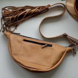 K&ouml;nnte beinhalten: Eine hellbraune Bauchtasche aus Leder in Halbmondform mit einer vorderen Rei&szlig;verschlusstasche mit schwarzem Rei&szlig;verschluss und einem langen, verstellbaren Riemen, der mit mehreren h&auml;ngenden Lederfransen an einem silberfarbenen Karabinerhaken versehen ist.
