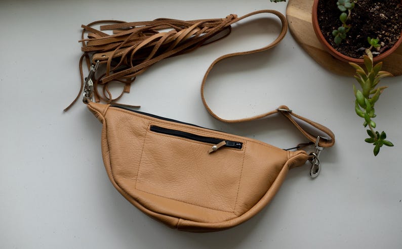 K&ouml;nnte beinhalten: Eine hellbraune Bauchtasche aus Leder in Halbmondform mit einer vorderen Rei&szlig;verschlusstasche mit schwarzem Rei&szlig;verschluss und einem langen, verstellbaren Riemen, der mit mehreren h&auml;ngenden Lederfransen an einem silberfarbenen Karabinerhaken versehen ist.