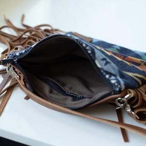 May include: A crossbody bag featuring a dark blue fabric panel with a multi-coloured geometric pattern, accented by brown leather trim and long fringe details. The bag has a black zip closure and a brown interior lining with a small zipped pocket.