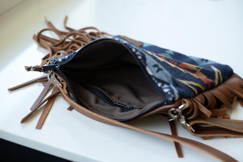 May include: A crossbody bag featuring a dark blue fabric panel with a multi-coloured geometric pattern, accented by brown leather trim and long fringe details. The bag has a black zip closure and a brown interior lining with a small zipped pocket.