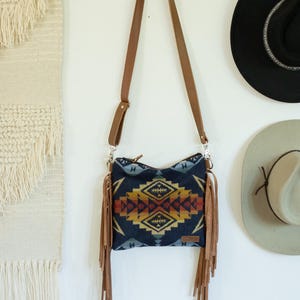 May include: A crossbody bag featuring a dark blue fabric body with a geometric pattern in shades of yellow, orange, and red. The bag includes long tan leather fringe on both sides, a matching tan leather adjustable shoulder strap, and a zip closure.