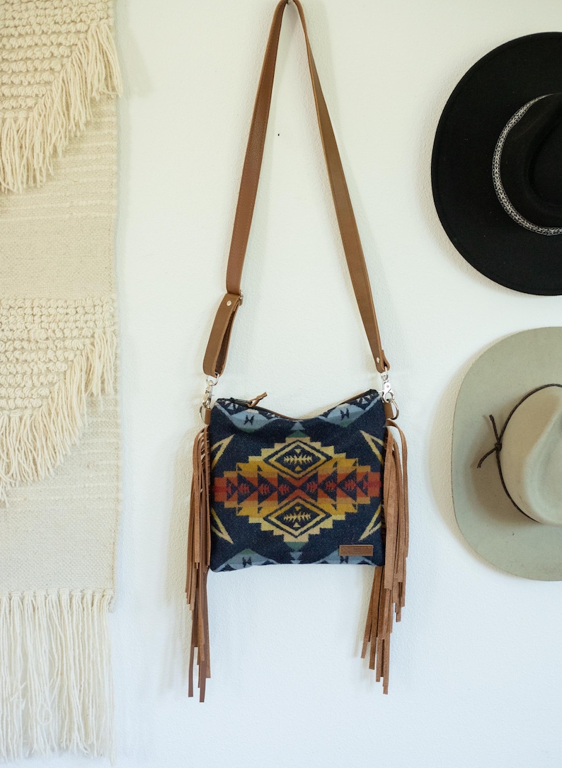 May include: A crossbody bag featuring a dark blue fabric body with a geometric pattern in shades of yellow, orange, and red. The bag includes long tan leather fringe on both sides, a matching tan leather adjustable shoulder strap, and a zip closure.