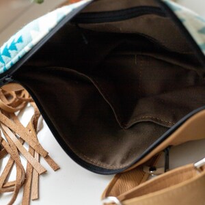 K&ouml;nnte beinhalten: Eine beigefarbene Umh&auml;ngetasche aus Leder mit einem Stoffeinsatz in T&uuml;rkis und Wei&szlig; mit geometrischem Muster. Die Tasche hat einen schwarzen Rei&szlig;verschluss, ein dunkelbraunes Innenfutter aus Stoff mit Einsteckfach und lange Lederfransen.