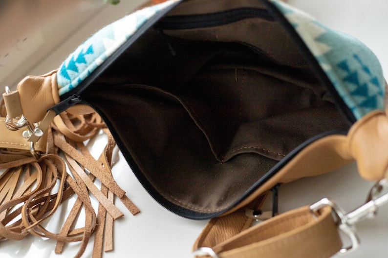 K&ouml;nnte beinhalten: Eine beigefarbene Umh&auml;ngetasche aus Leder mit einem Stoffeinsatz in T&uuml;rkis und Wei&szlig; mit geometrischem Muster. Die Tasche hat einen schwarzen Rei&szlig;verschluss, ein dunkelbraunes Innenfutter aus Stoff mit Einsteckfach und lange Lederfransen.