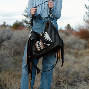 Puede incluir: Un bolso hobo negro con largos flecos en negro y beige. El bolso incluye un panel central con un patr&oacute;n geom&eacute;trico en negro, blanco y beige. Est&aacute; equipado con una correa de hombro negra sujeta mediante herrajes de metal de tono plateado.