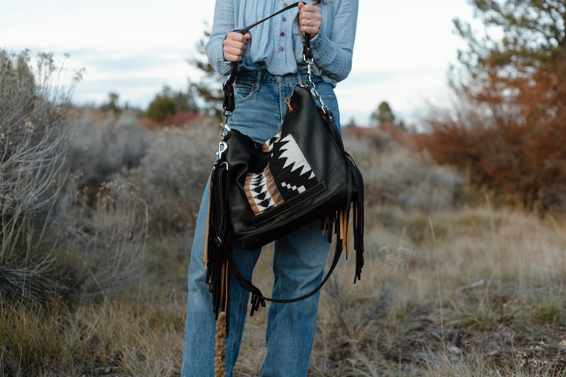 Puede incluir: Un bolso hobo negro con largos flecos en negro y beige. El bolso incluye un panel central con un patr&oacute;n geom&eacute;trico en negro, blanco y beige. Est&aacute; equipado con una correa de hombro negra sujeta mediante herrajes de metal de tono plateado.