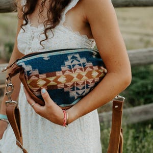 May include: A crossbody bag featuring a woven fabric panel with a geometric pattern in shades of blue, brown, cream, and orange. The bag includes brown leather sides and a matching brown leather adjustable strap attached with silver metal hardware.