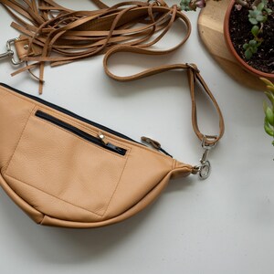 May include: A tan leather fanny pack featuring a crescent shape, a front zippered pocket with a black zipper, and a long adjustable strap adorned with multiple thin leather fringe tassels attached to a silver metal swivel clasp on the side.