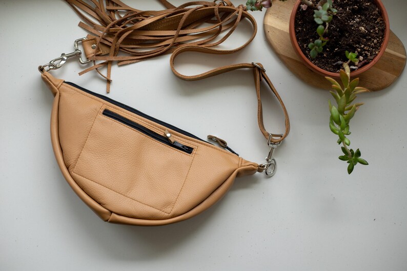 May include: A tan leather fanny pack featuring a crescent shape, a front zippered pocket with a black zipper, and a long adjustable strap adorned with multiple thin leather fringe tassels attached to a silver metal swivel clasp on the side.