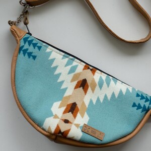 K&ouml;nnte beinhalten: Eine halbmondf&ouml;rmige Umh&auml;ngetasche mit einem hellblauen Stoffeinsatz, der ein geometrisches Muster in Wei&szlig;, Beige und Dunkelbraun aufweist. Die Tasche ist mit beigefarbenem Leder eingefasst und hat einen passenden Riemen mit langen Fransen.