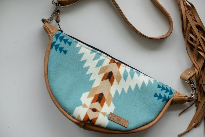 K&ouml;nnte beinhalten: Eine halbmondf&ouml;rmige Umh&auml;ngetasche mit einem hellblauen Stoffeinsatz, der ein geometrisches Muster in Wei&szlig;, Beige und Dunkelbraun aufweist. Die Tasche ist mit beigefarbenem Leder eingefasst und hat einen passenden Riemen mit langen Fransen.