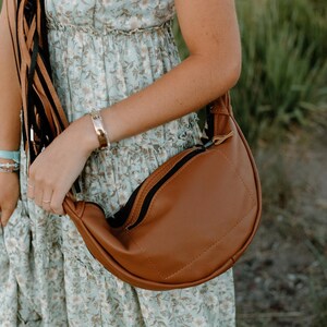 May include: A crescent-shaped brown crossbody bag with a smooth finish and a black zipper closure. The bag features a long strap adorned with multiple thin, dangling black and brown leather-like fringe tassels hanging from the side attachment point.