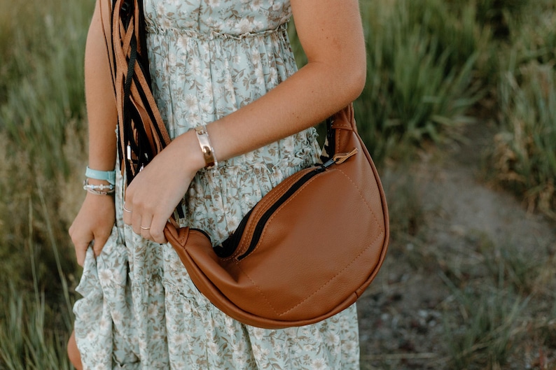 May include: A crescent-shaped brown crossbody bag with a smooth finish and a black zipper closure. The bag features a long strap adorned with multiple thin, dangling black and brown leather-like fringe tassels hanging from the side attachment point.