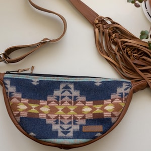 May include: A crescent-shaped fanny pack featuring a blue, brown, and lime green geometric pattern on the front panel. The bag has brown leather trim, a matching adjustable strap, and a long fringe detail attached to the side of the bag.