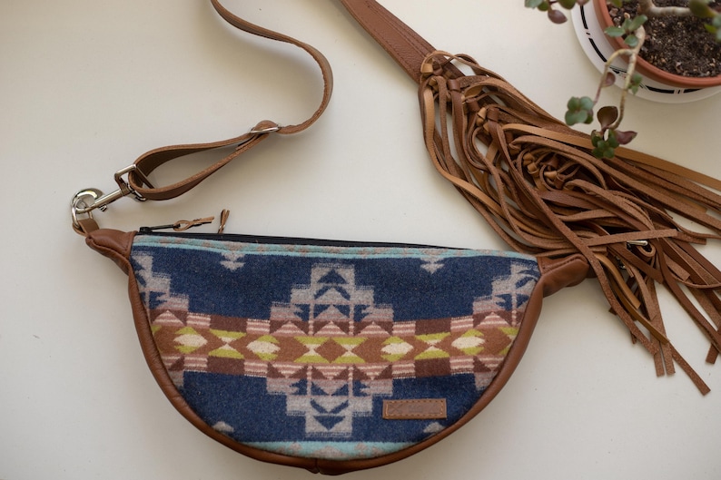 May include: A crescent-shaped fanny pack featuring a blue, brown, and lime green geometric pattern on the front panel. The bag has brown leather trim, a matching adjustable strap, and a long fringe detail attached to the side of the bag.
