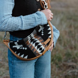 May include: A crescent-shaped crossbody bag featuring a woven textile pattern in black, white, and tan. The bag is trimmed with tan leather piping and includes a matching leather strap with a metal swivel clip attached to a metal D-ring.