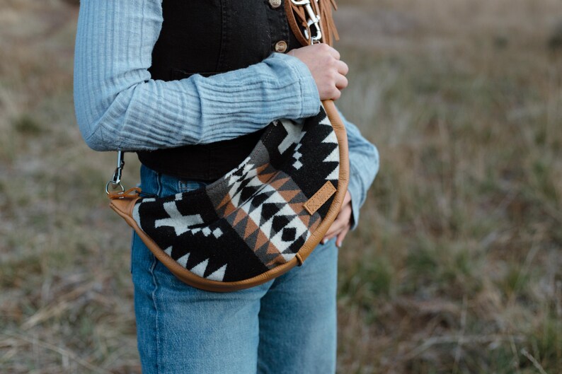 May include: A crescent-shaped crossbody bag featuring a woven textile pattern in black, white, and tan. The bag is trimmed with tan leather piping and includes a matching leather strap with a metal swivel clip attached to a metal D-ring.