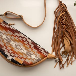 May include: A half-moon shaped bag with a Southwestern pattern in shades of brown, orange, and cream. The bag has a leather trim and a long, adjustable strap. A separate leather fringe detail is also included.