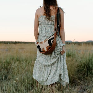 May include: A light green floral print tiered maxi dress with spaghetti straps paired with a crescent-shaped crossbody bag. The bag features a tan leather base, a cream, black, and brown geometric patterned fabric panel, and long black leather fringe.
