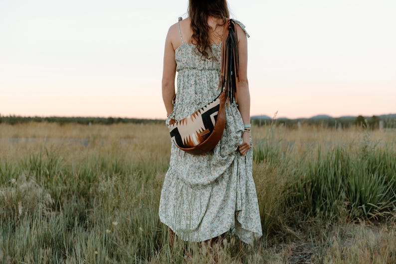 May include: A light green floral print tiered maxi dress with spaghetti straps paired with a crescent-shaped crossbody bag. The bag features a tan leather base, a cream, black, and brown geometric patterned fabric panel, and long black leather fringe.
