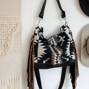 Puede incluir: Un bolso de hombro estilo hobo con un patr&oacute;n geom&eacute;trico en negro, blanco y tostado en el cuerpo principal. El bolso incluye una base de cuero negro s&oacute;lido, una correa ajustable negra con herrajes plateados y largos flecos en tonos tostado y negro.