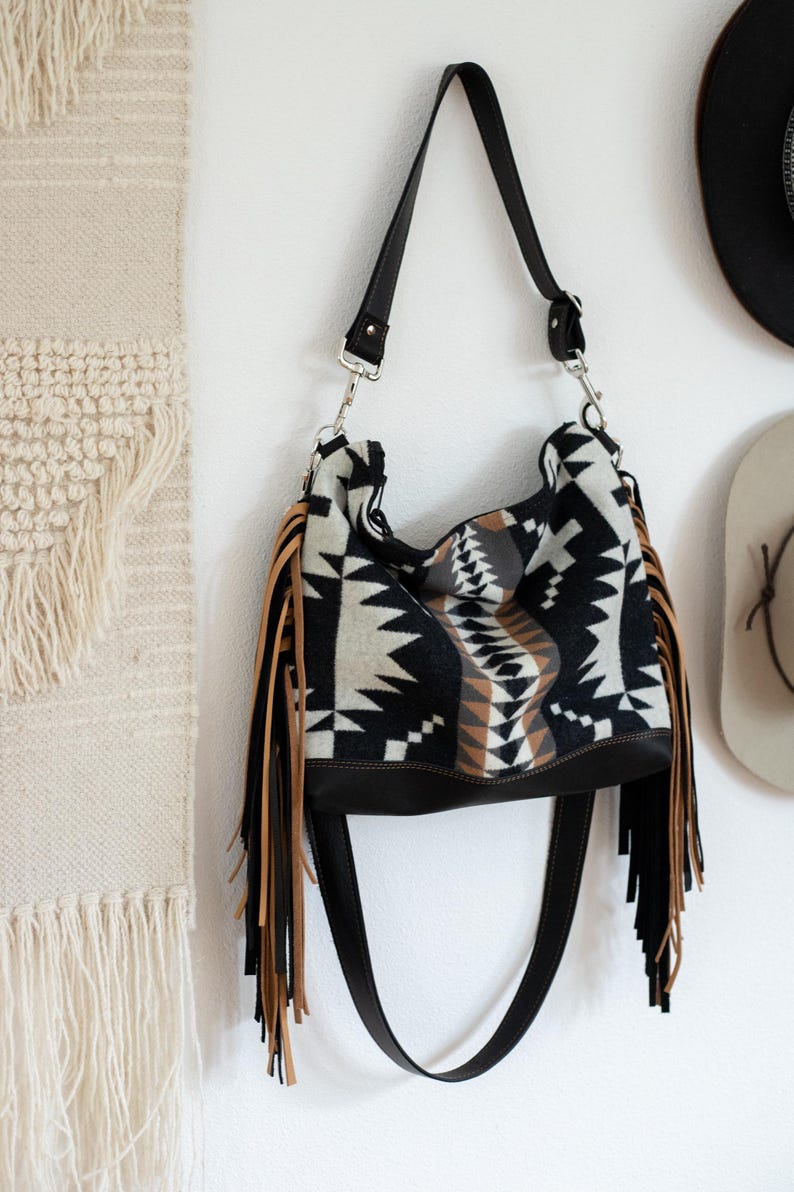 Puede incluir: Un bolso de hombro estilo hobo con un patr&oacute;n geom&eacute;trico en negro, blanco y tostado en el cuerpo principal. El bolso incluye una base de cuero negro s&oacute;lido, una correa ajustable negra con herrajes plateados y largos flecos en tonos tostado y negro.