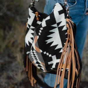 Puede incluir: Un bolso de hombro estilo hobo con un patr&oacute;n geom&eacute;trico en negro, blanco y tostado. El bolso est&aacute; decorado con largos flecos de cuero color tostado claro en los laterales e incluye una correa de cuero marr&oacute;n oscuro con herrajes met&aacute;licos plateados.