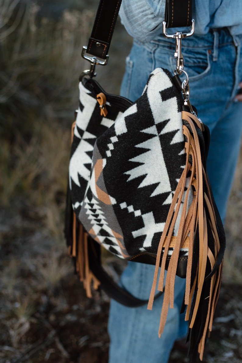 Puede incluir: Un bolso de hombro estilo hobo con un patr&oacute;n geom&eacute;trico en negro, blanco y tostado. El bolso est&aacute; decorado con largos flecos de cuero color tostado claro en los laterales e incluye una correa de cuero marr&oacute;n oscuro con herrajes met&aacute;licos plateados.