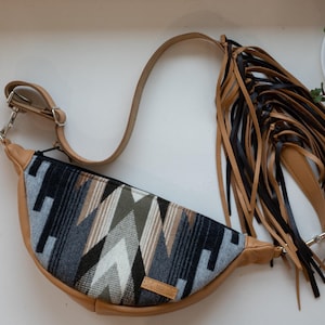 May include: A tan leather fanny pack with a geometric patterned front in shades of gray, black, and olive green. The bag has a long adjustable strap and a decorative fringe of black and tan leather strips. The bag is approximately 12 inches wide.