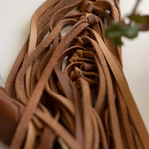 May include: A collection of tan and brown leather strips knotted together to form a dense, layered fringe. The strips vary in shade and texture, creating a complex, intertwined structure with a smooth, matte finish and visible grain patterns.