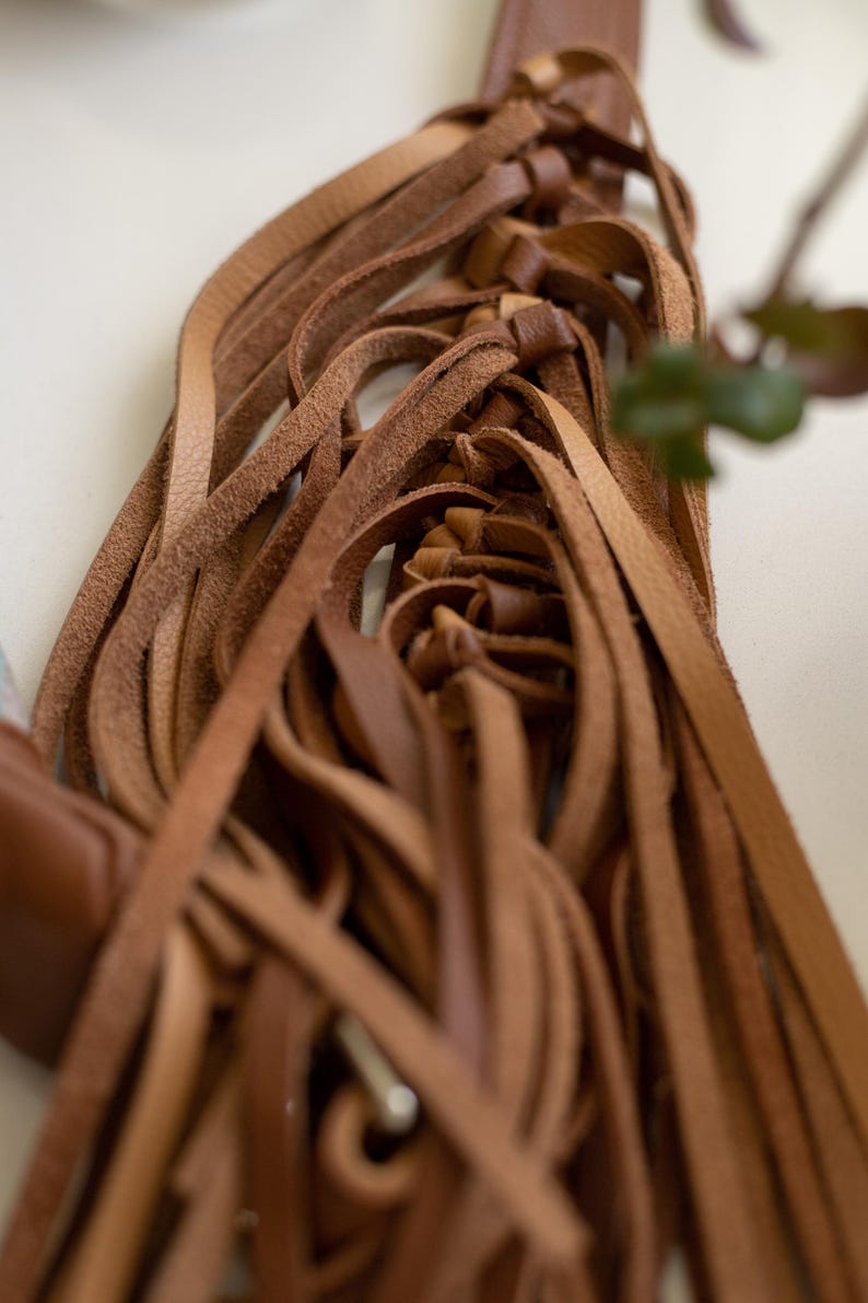 May include: A collection of tan and brown leather strips knotted together to form a dense, layered fringe. The strips vary in shade and texture, creating a complex, intertwined structure with a smooth, matte finish and visible grain patterns.