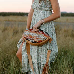 May include: A crescent-shaped crossbody bag featuring a multi-colored geometric pattern in shades of orange, brown, and cream on the upper panel, paired with a solid tan leather base, an adjustable tan leather strap, and decorative leather fringe.