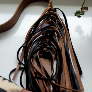 May include: A bag featuring a long brown leather strap and a decorative cluster of dangling leather fringe strips in alternating black and tan colors. The fringe is attached to a brown leather base, with a portion of a patterned fabric bag visible.