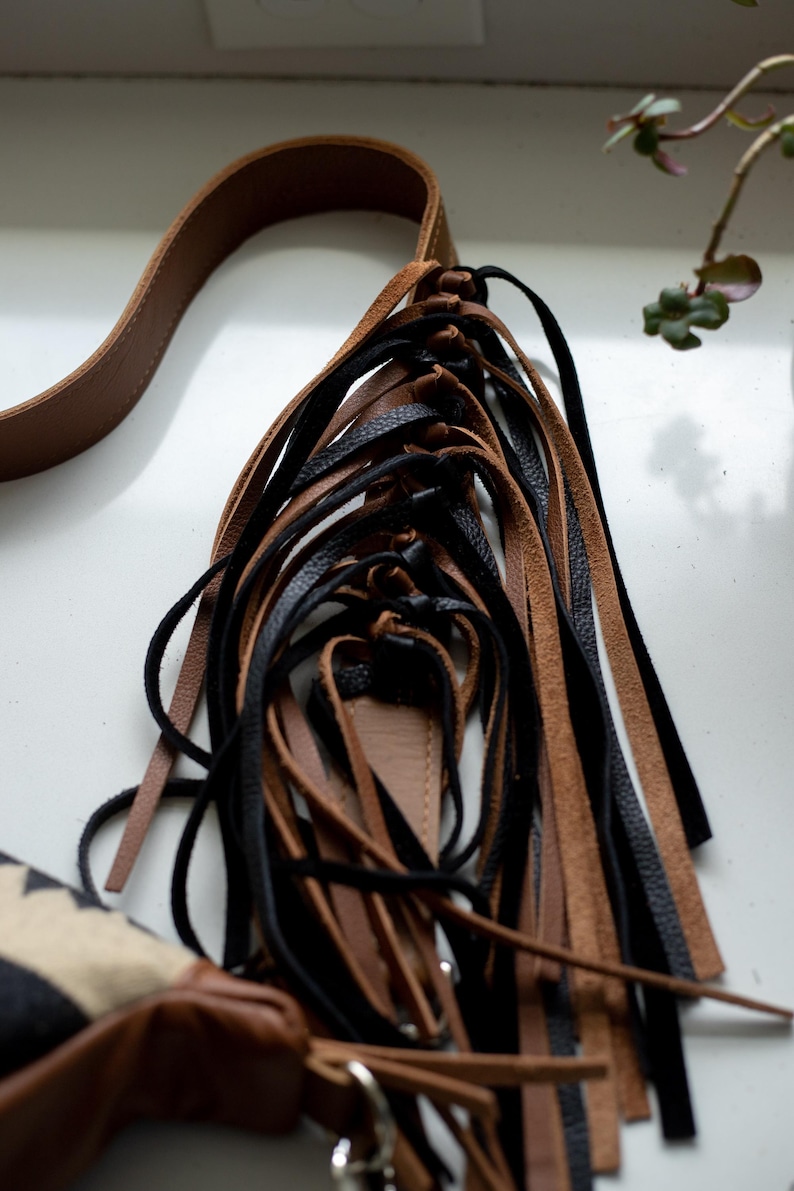 May include: A bag featuring a long brown leather strap and a decorative cluster of dangling leather fringe strips in alternating black and tan colors. The fringe is attached to a brown leather base, with a portion of a patterned fabric bag visible.