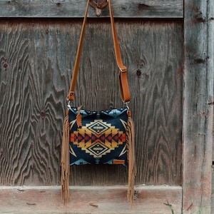May include: A crossbody bag featuring a dark blue fabric body with a geometric pattern in shades of yellow, orange, and red. The bag includes a long tan leather strap and long tan leather fringe detailing hanging from both side edges of the bag.
