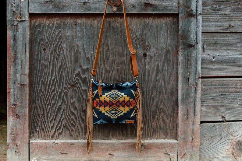 May include: A crossbody bag featuring a dark blue fabric body with a geometric pattern in shades of yellow, orange, and red. The bag includes a long tan leather strap and long tan leather fringe detailing hanging from both side edges of the bag.