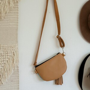 May include: A tan crossbody bag with a smooth finish, a black zipper closure, and a matching tan leather tassel attached to the side. The bag features a long, adjustable shoulder strap with silver-toned metal hardware and a curved bottom edge.