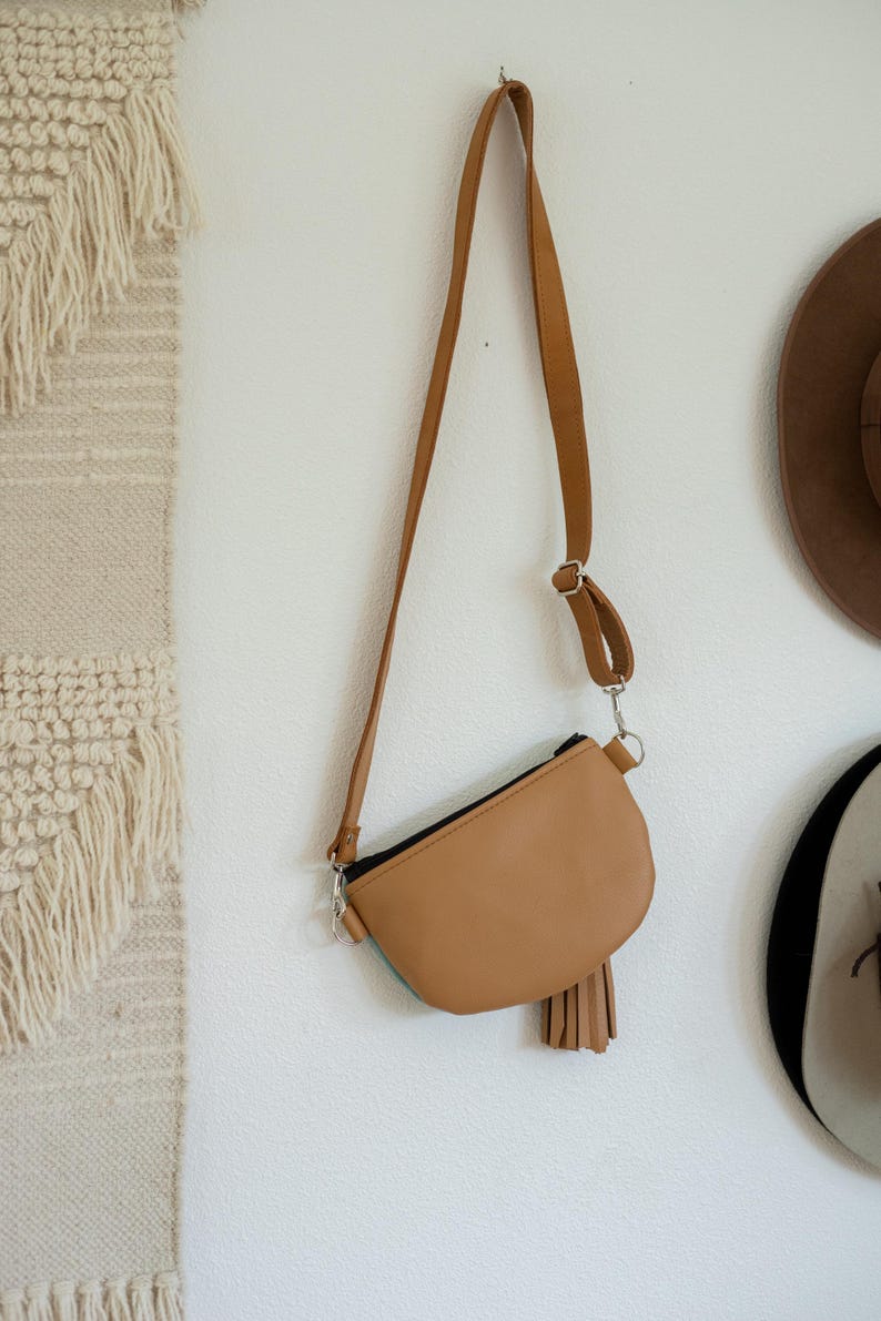 May include: A tan crossbody bag with a smooth finish, a black zipper closure, and a matching tan leather tassel attached to the side. The bag features a long, adjustable shoulder strap with silver-toned metal hardware and a curved bottom edge.
